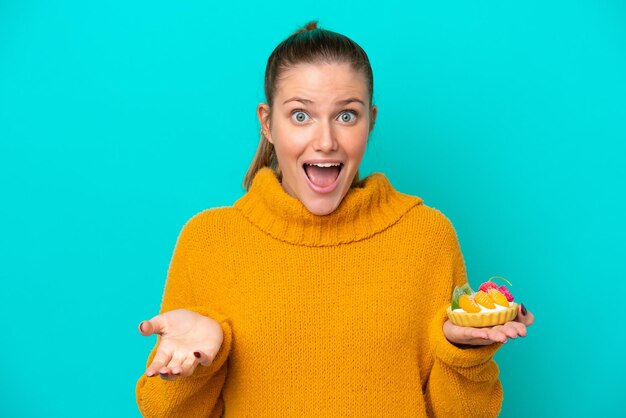 Young caucasian woman holding a tartlet isolated on blue background with shocked facial expression