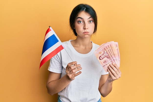 Young caucasian woman holding thailand flag and baht banknotes puffing cheeks with funny face. mouth inflated with air, catching air.