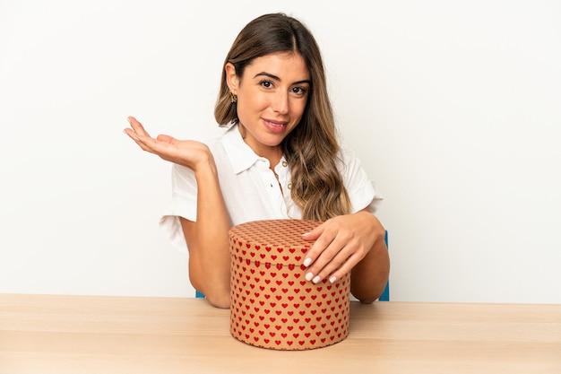 Young caucasian woman holding a valentines day box isolated showing a copy space on a palm and holding another hand on waist.