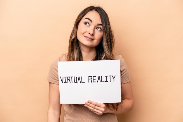 Young caucasian woman holding a virtual reality placard isolated on beige background dreaming of achieving goals and purposes