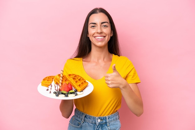 Young caucasian woman holding waffles isolated on pink background giving a thumbs up gesture