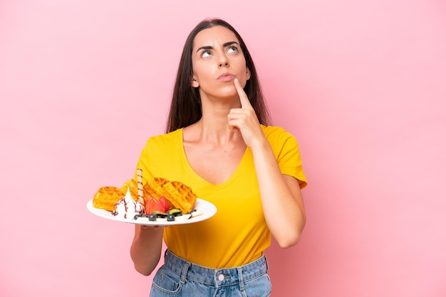 Young caucasian woman holding waffles isolated on pink background having doubts while looking up