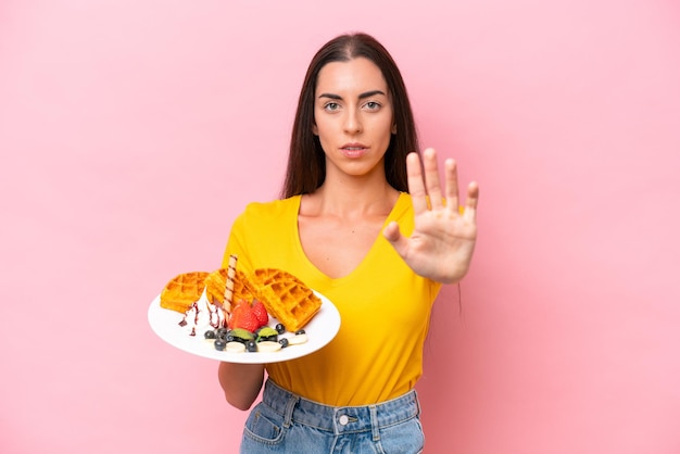 Young caucasian woman holding waffles isolated on pink background making stop gesture