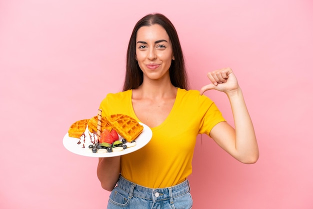 Young caucasian woman holding waffles isolated on pink background proud and selfsatisfied