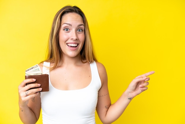 Young caucasian woman holding a wallet isolated on yellow background surprised and pointing finger to the side