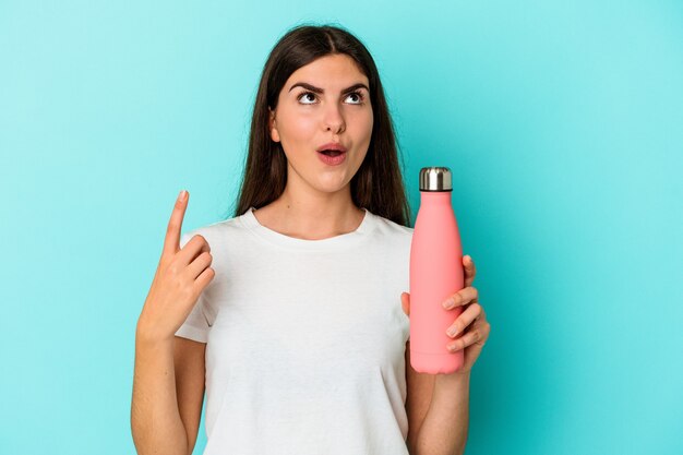 Young caucasian woman holding water bottle isolated on blue wall pointing upside with opened mouth