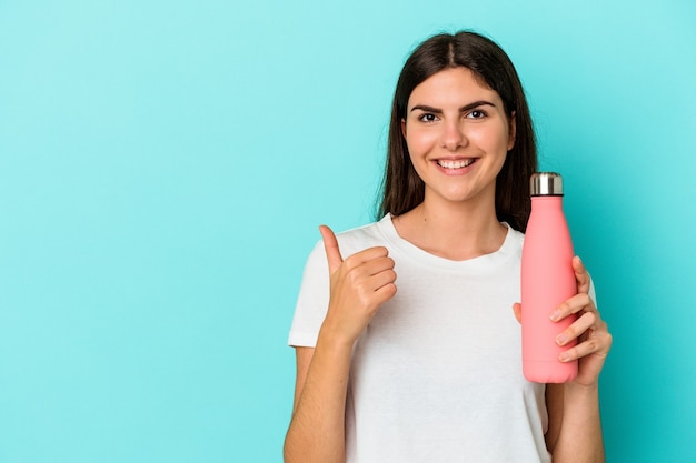 Young caucasian woman holding water bottle isolated on blue wall smiling and raising thumb up