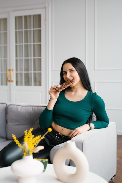 Young Caucasian woman holds a chocolate bar in her hand and eats it while sitting on the couch at home