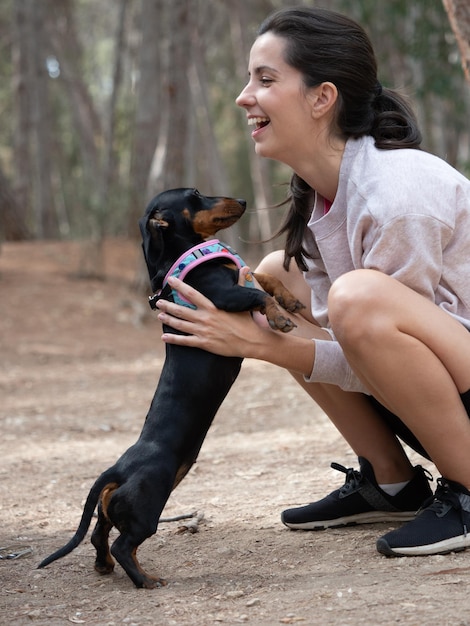 Young caucasian woman hugging black smooth haired miniature dachshund in forest