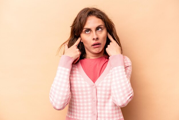 Young caucasian woman isolated on beige background covering ears with fingers stressed and desperate by a loudly ambient