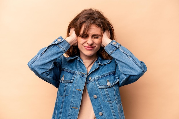 Young caucasian woman isolated on beige background covering ears with hands