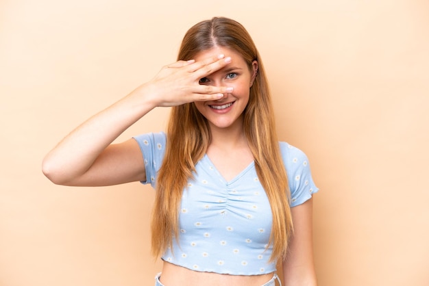 Young caucasian woman isolated on beige background covering eyes by hands and smiling