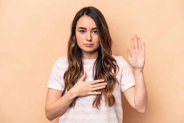 Young caucasian woman isolated on beige background taking an oath putting hand on chest