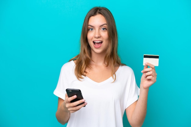 Young caucasian woman isolated on blue background buying with the mobile and holding a credit card with surprised expression