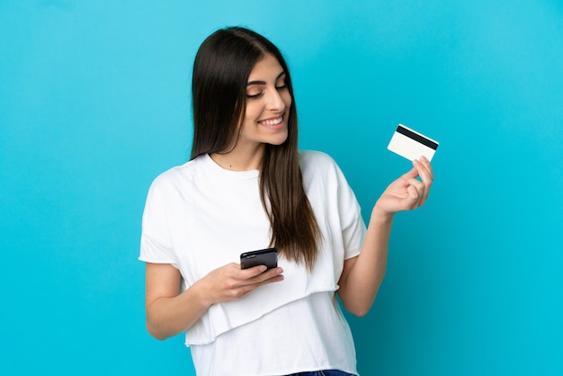 Young caucasian woman isolated on blue background buying with the mobile with a credit card while thinking