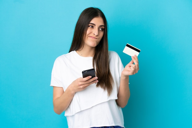 Young caucasian woman isolated on blue background buying with the mobile with a credit card while thinking