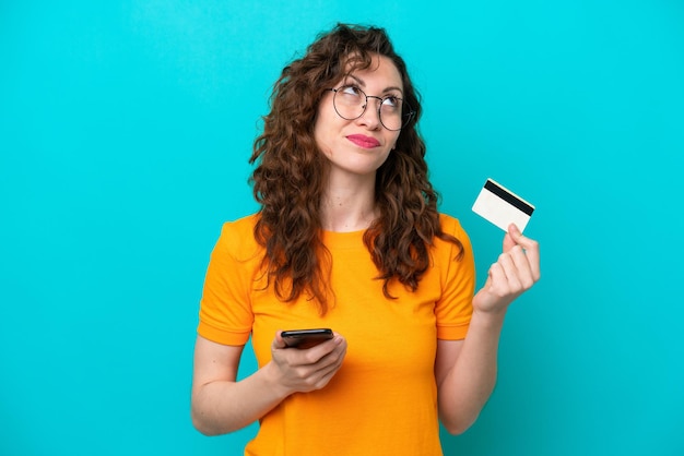 Young caucasian woman isolated on blue background buying with the mobile with a credit card while thinking