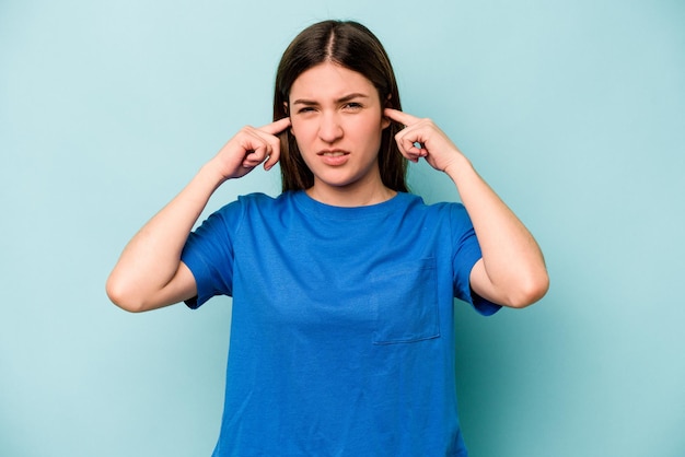 Young caucasian woman isolated on blue background covering ears with fingers stressed and desperate by a loudly ambient