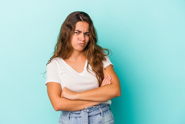 Young caucasian woman isolated on blue background  frowning face in displeasure, keeps arms folded.