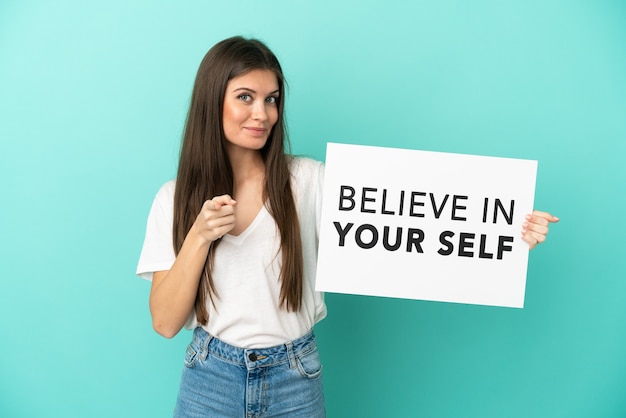 Young caucasian woman isolated on blue background holding a placard with text Believe In Your Self and pointing to the front