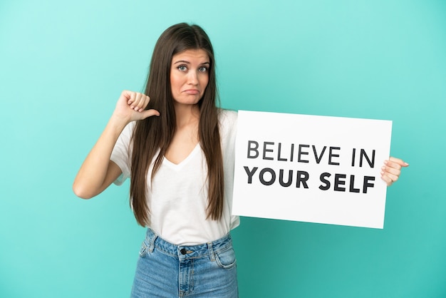 Young caucasian woman isolated on blue background holding a placard with text Believe In Your Self with proud gesture