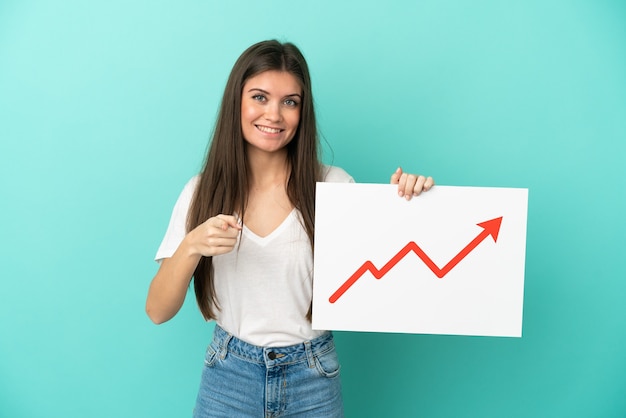 Young caucasian woman isolated on blue background holding a sign with a growing statistics arrow symbol and pointing to the front