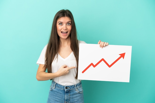 Young caucasian woman isolated on blue background holding a sign with a growing statistics arrow symbol with surprised expression