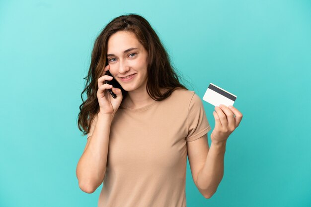 Young caucasian woman isolated on blue background keeping a conversation with the mobile phone and holding a credit card