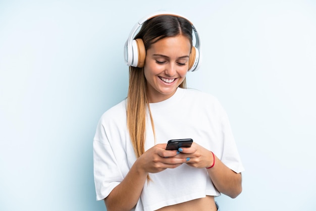 Young caucasian woman isolated on blue background listening music and looking to mobile