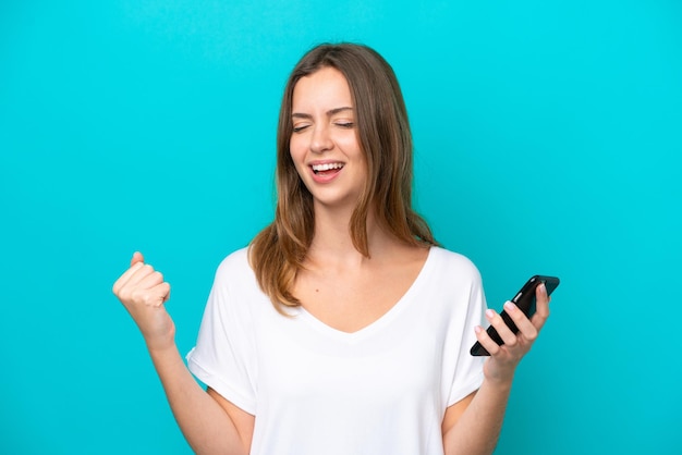 Young caucasian woman isolated on blue background using mobile phone and doing victory gesture