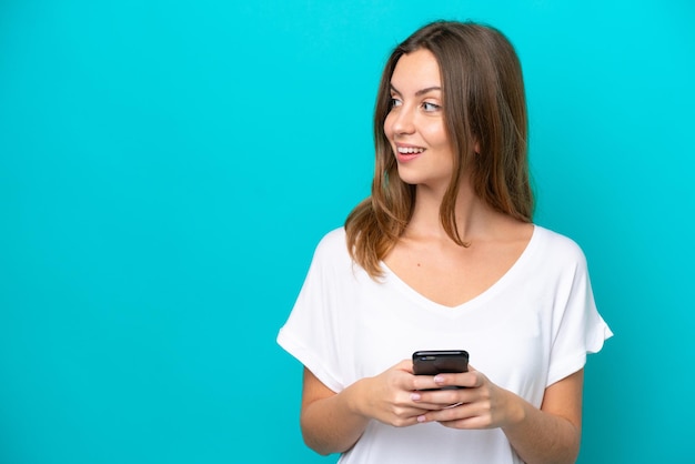 Young caucasian woman isolated on blue background using mobile phone and looking up