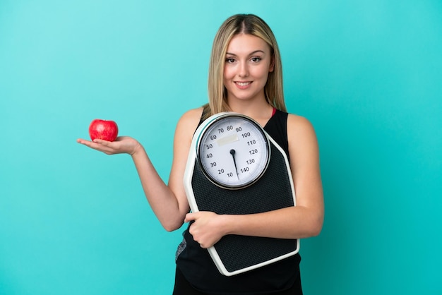 Young caucasian woman isolated on blue background with weighing machine and with an apple