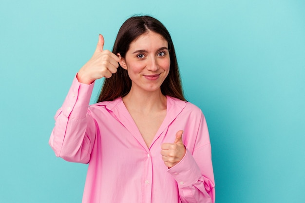 Young caucasian woman isolated on blue wall raising both thumbs up, smiling and confident.