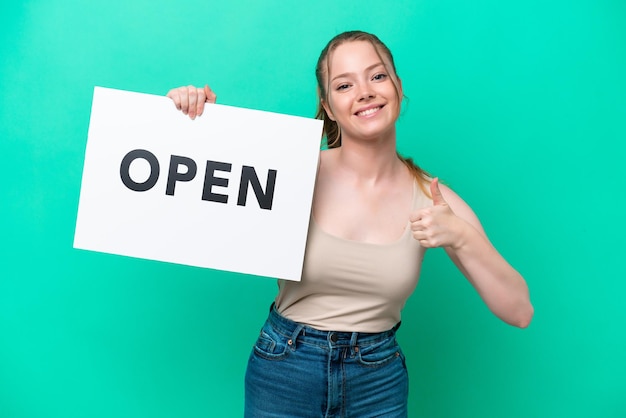 Young caucasian woman isolated on green background holding a placard with text OPEN with thumb up