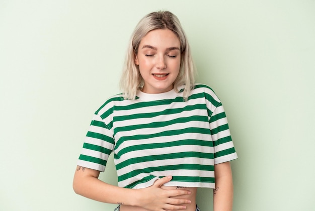 Young caucasian woman isolated on green background touches tummy, smiles gently, eating and satisfaction concept.