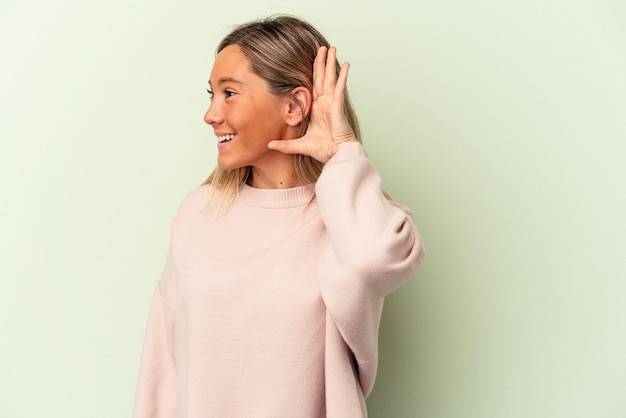 Young caucasian woman isolated on green background trying to listening a gossip.