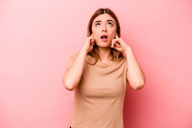 Young caucasian woman isolated on pink background covering ears with fingers stressed and desperate by a loudly ambient