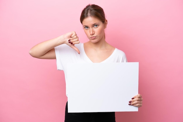 Young caucasian woman isolated on pink background holding an empty placard and doing bad signal