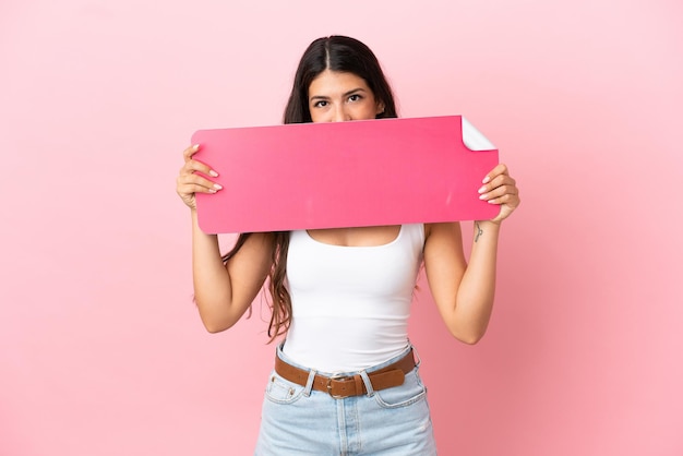 Young caucasian woman isolated on pink background holding an empty placard and hiding behind it