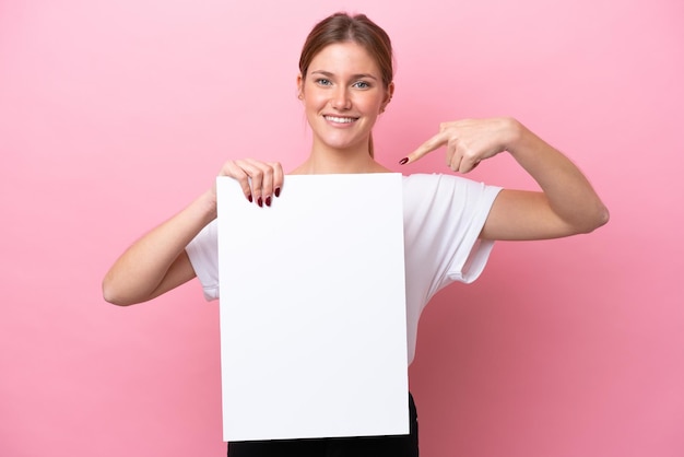 Young caucasian woman isolated on pink background holding an empty placard with happy expression and pointing it