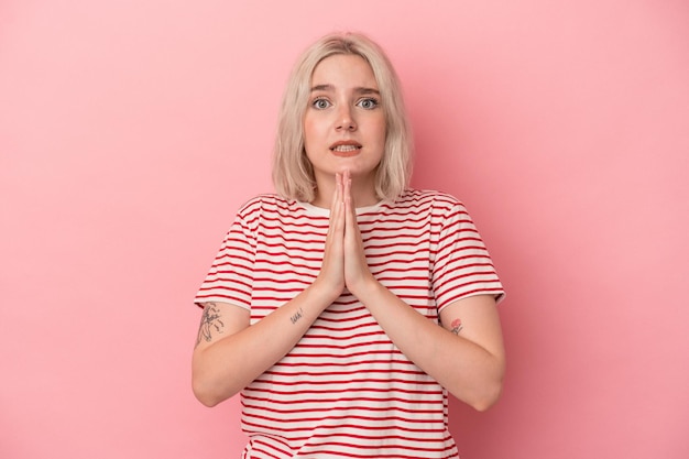 Young caucasian woman isolated on pink background holding hands in pray near mouth feels confident