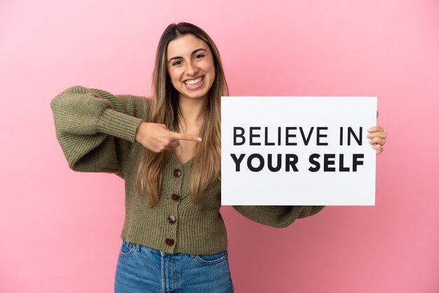 Young caucasian woman isolated on pink background holding a placard with text Believe In Your Self and pointing it