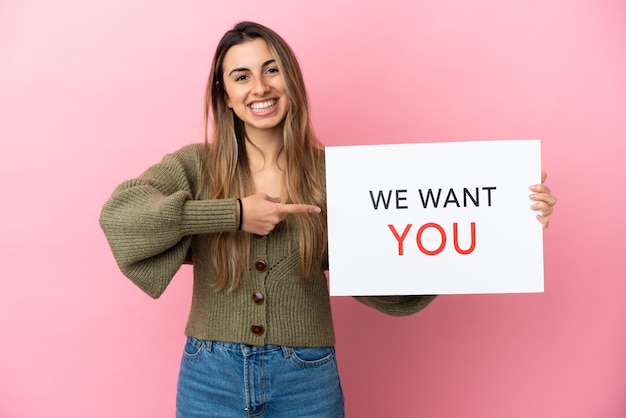 Young caucasian woman isolated on pink background holding We Want You board and  pointing it