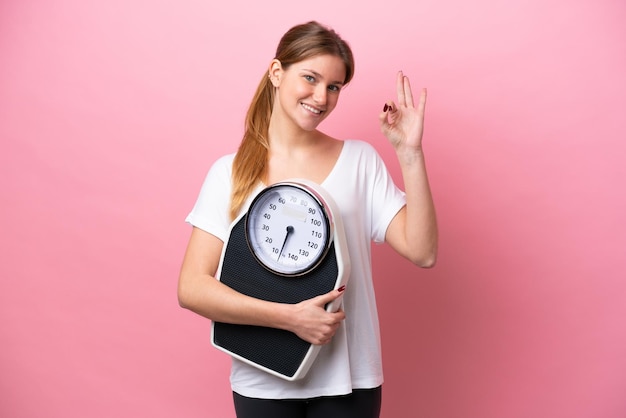 Young caucasian woman isolated on pink background holding a weighing machine and doing OK sign