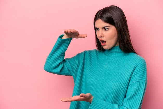 Young caucasian woman isolated on pink background shocked and amazed holding a copy space between hands