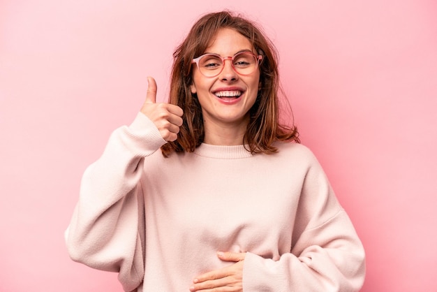 Young caucasian woman isolated on pink background touches tummy smiles gently eating and satisfaction concept