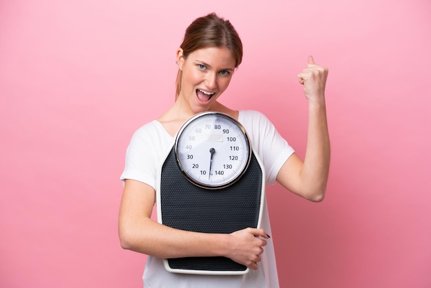 Young caucasian woman isolated on pink background with weighing machine and doing victory gesture