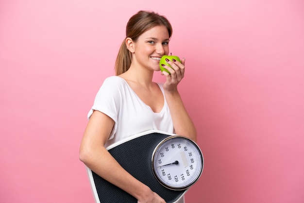 Young caucasian woman isolated on pink background with weighing machine and with an apple