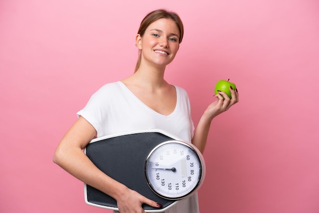 Young caucasian woman isolated on pink background with weighing machine and with an apple