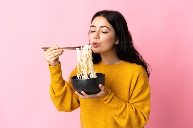 Young caucasian woman isolated on pink wall holding a bowl of noodles with chopstick sand blowing it because they are hot